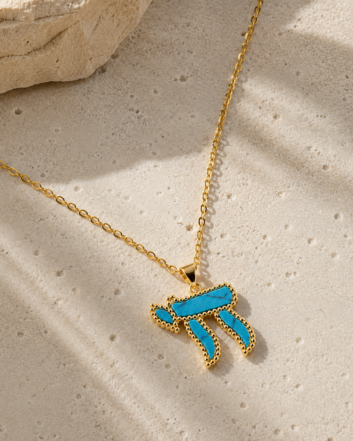 Gold tone Perlée Chai Necklace with a turquoise Chai pendant styled on a neutral stone surface.