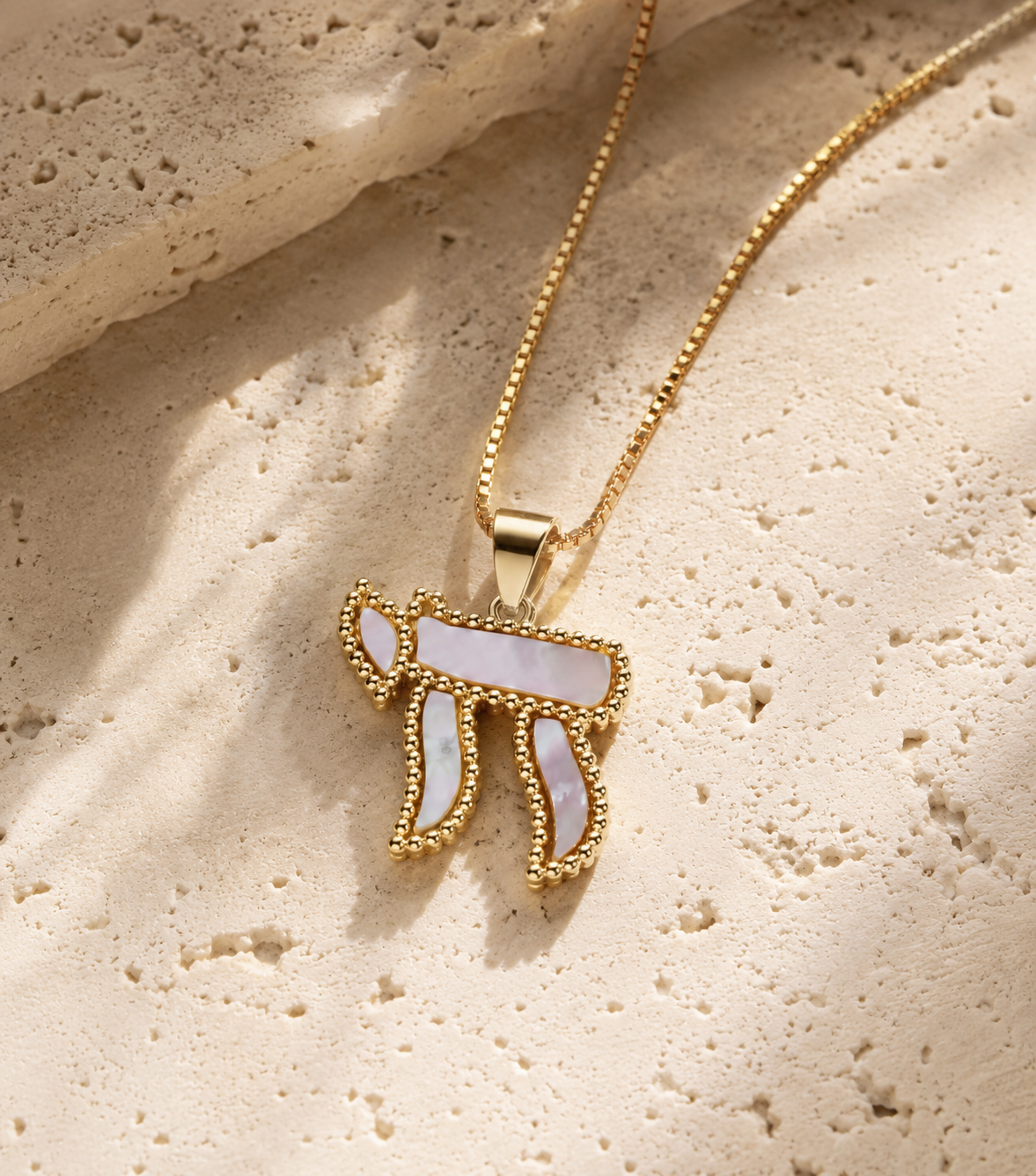 Gold tone Perlée Chai Necklace with white pearl-effect detailing styled on a neutral stone surface.