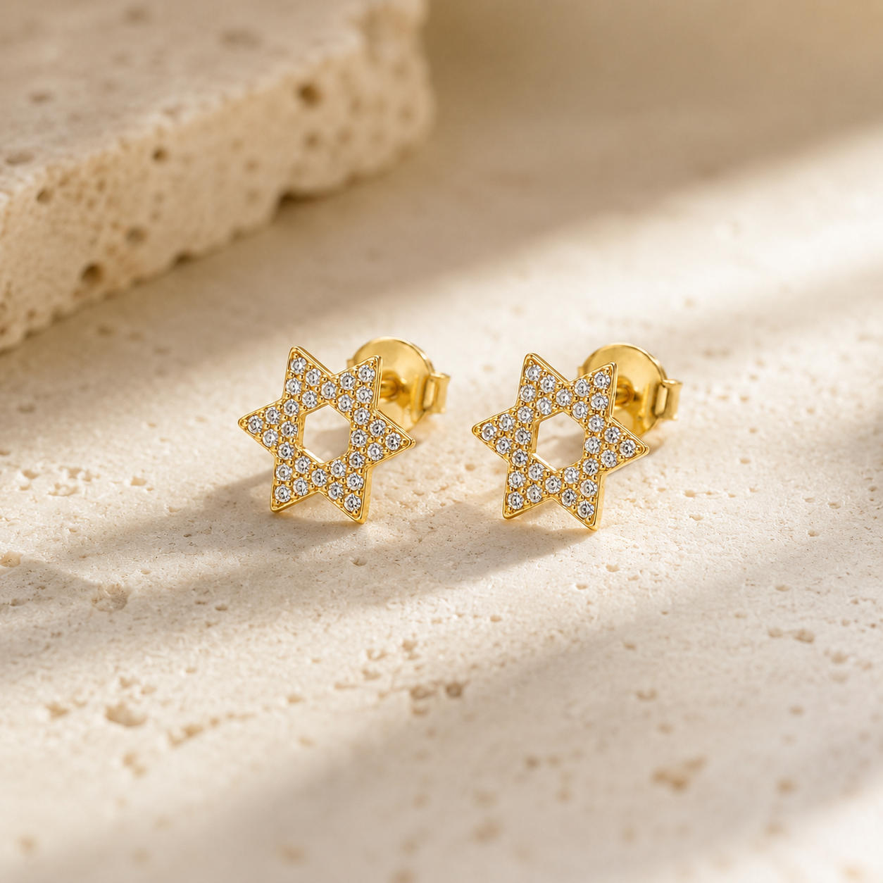 Gold tone Magen David pavé stud earrings styled on a neutral stone surface.