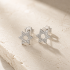 Silver tone Magen David pavé stud earrings styled on a neutral stone surface.
