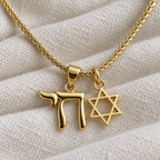 Close-up of the Neshama necklace's gold tone Chai and open Star of David charms on ivory linen.