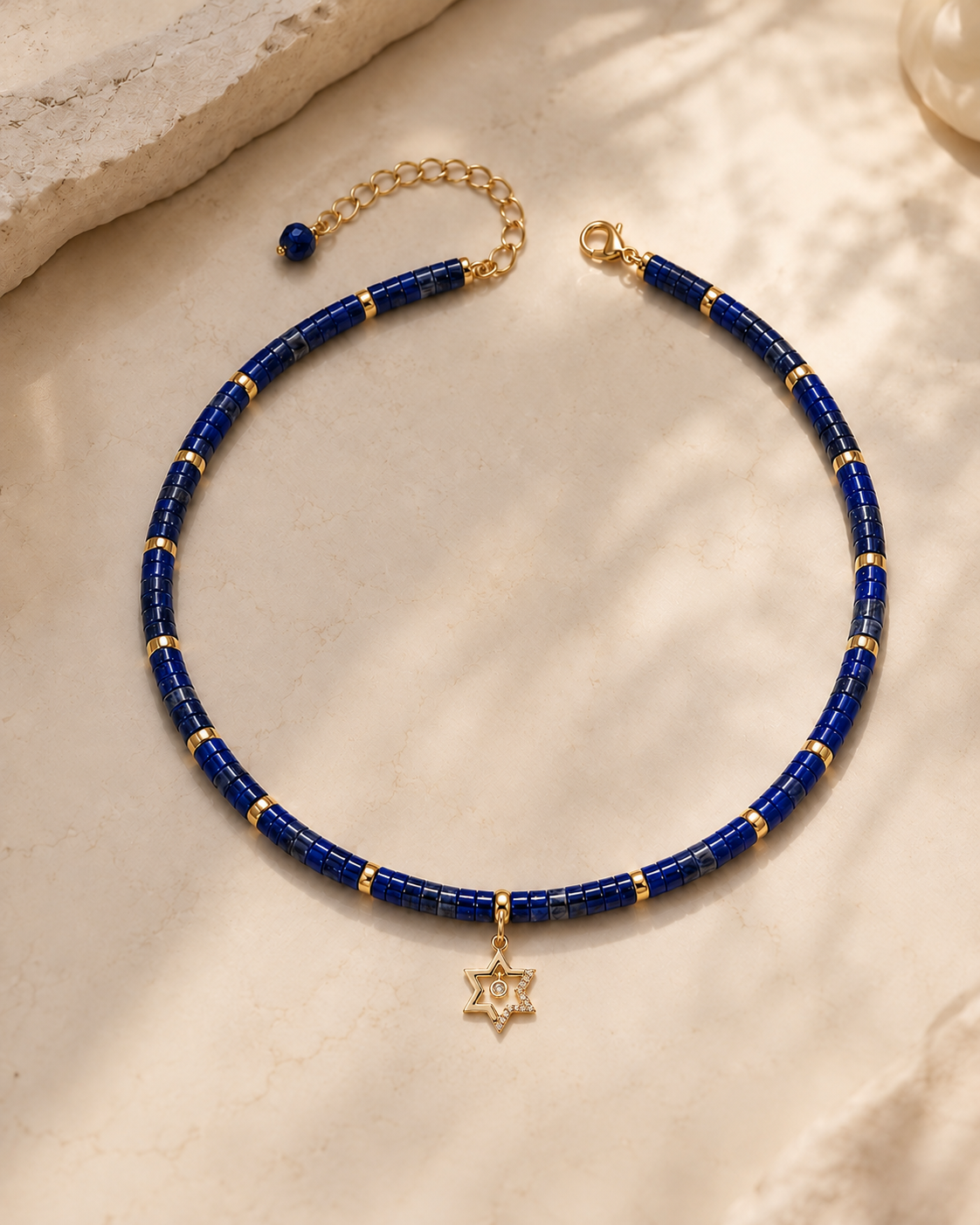Noa Star of David Beaded Choker Necklace in Lapis with gold tone details on neutral stone surface.