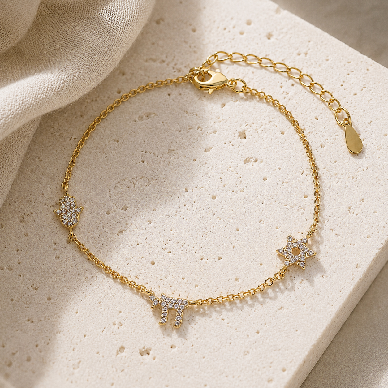 Gold Tone Pavé Trio Bracelet with Star of David, Hamsa, and Chai charms on a luxury neutral stone surface.